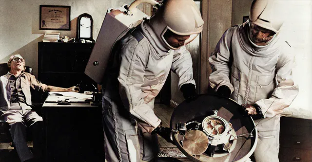 Two scientists in space suits examine a space capsule. In the background, the body of a man sitting on a chair in front of a desk.