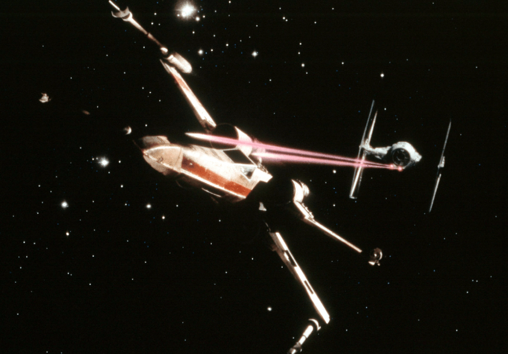 A TIE figther pursues an X-Wing starfighter and fires a double LASER salvo.